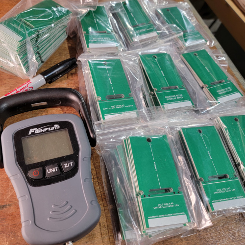 A Fishfun brand digital fishing scale lays on a workbench beside stacks of business card sized circuit boards