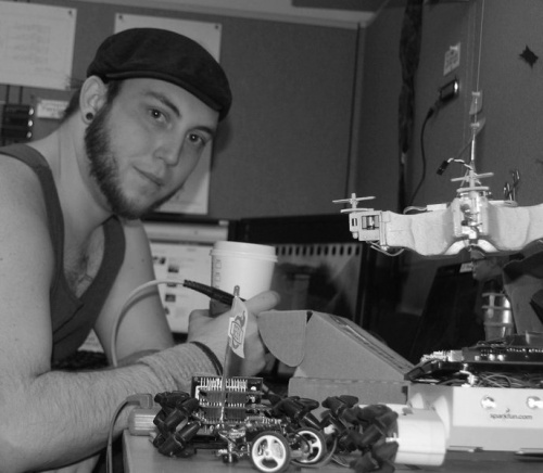 Hyperedge- , PCB Design, Hardware Design, Electronics Design, black and white, low-res photo of me sitting at a desk with several small robots on the desk in front of me. I'm holding a soldering iron and a cup of coffee. I'm wearing a tank top and a flat cap.