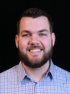 Professional headshot of a smiling bearded man wearing a light blue checkered button-down shirt against a black background